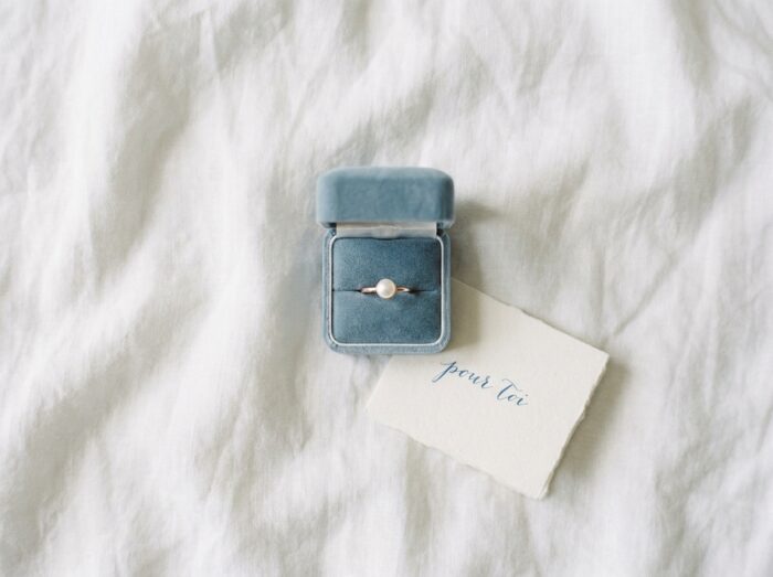 Écrin en velours bleu ouvert sur un drap blanc contenant une bague en or rose et perle avec un mot manuscrit, cadeau bijou romantique élégant