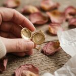 Mains féminines tenant un médaillon en or gravé d'un cœur, posé sur une table en bois avec des pétales de rose séchés, bijou sentiment romantique
